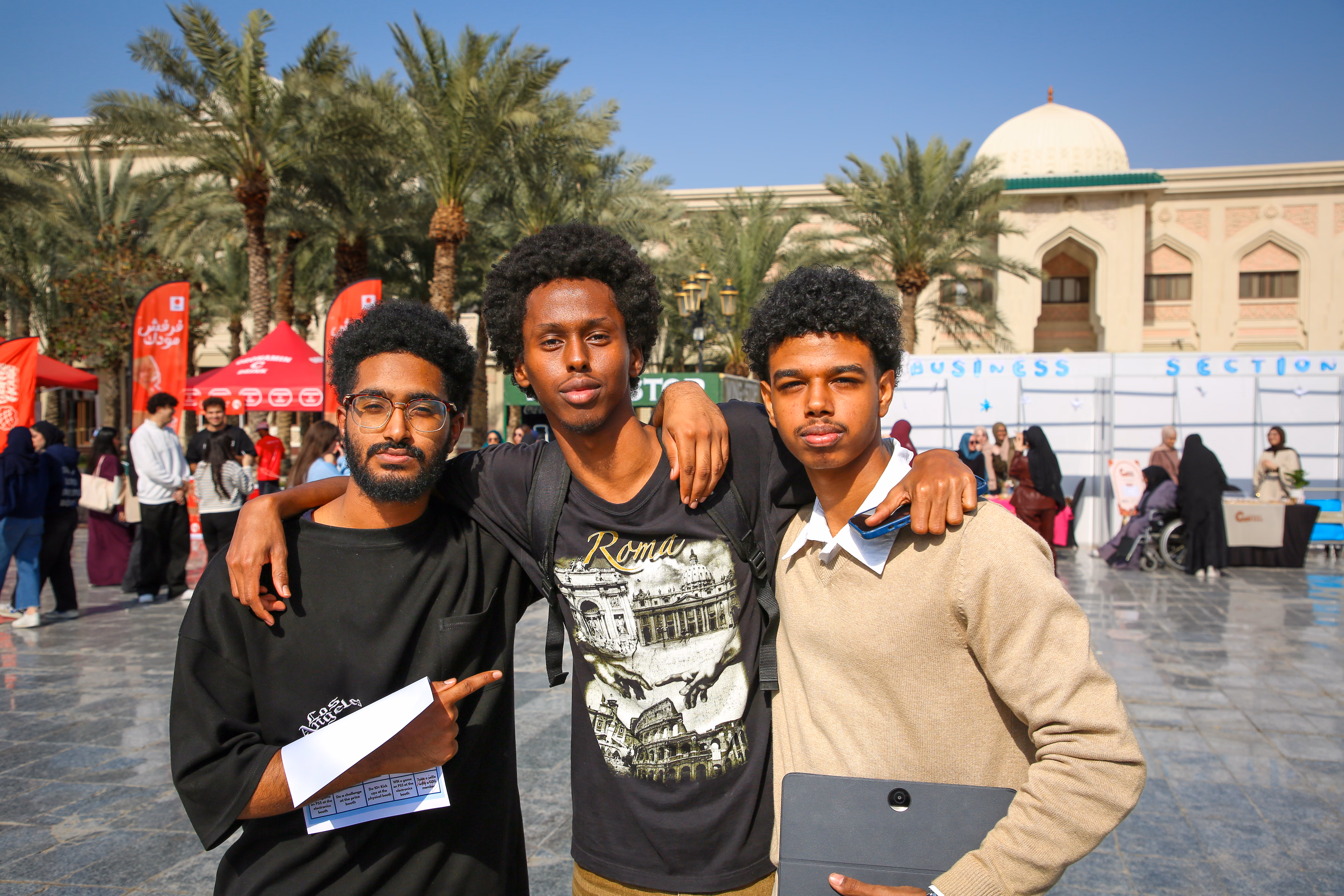 Three members arm in arm in the campus plaza, one holding a stack of flyers, with the fair's section booths behind them.